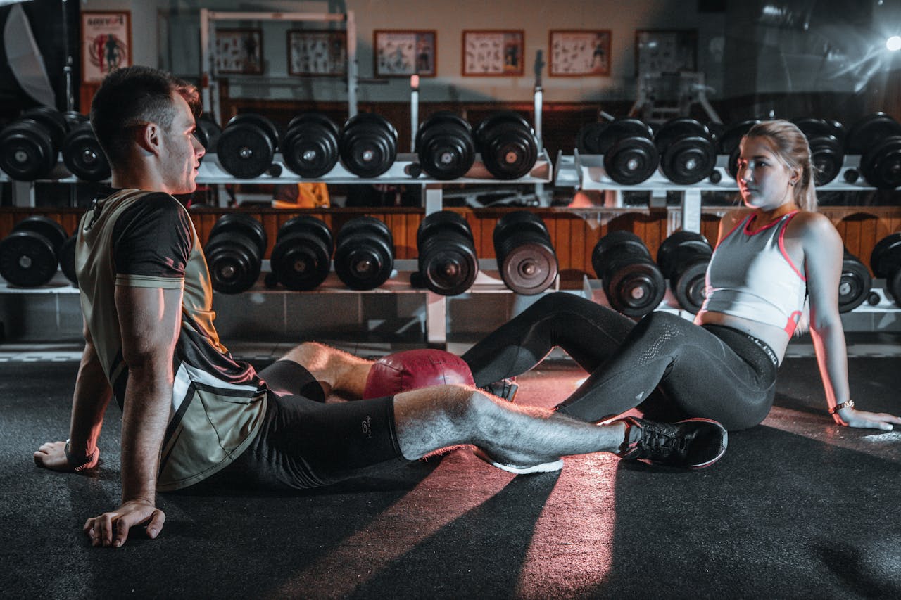 home-hero A man and woman resting together on the gym floor, surrounded by dumbbells.