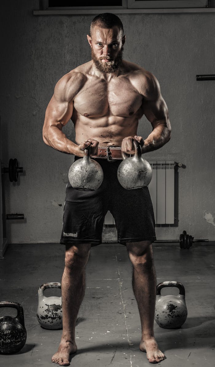 services-06 Shirtless man with muscular build lifting kettlebells indoors, showcasing strength and fitness.